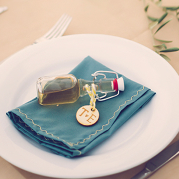 Petite Fiolle fermée par un bouchon avec joint orangé contenant une boisson, avec médaillon en bois personnalisé avec initiales, posé sur une serviette dans une assiette de table de mariage